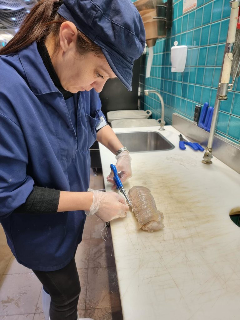 Apprentie en poissonnerie découpant soigneusement un produit de la mer sur une planche de travail propre, équipée de gants de protection et de ciseaux de cuisine. Elle porte une blouse et une casquette bleues, dans un environnement de cuisine professionnelle avec un mur en carrelage bleu et un évier en arrière-plan.